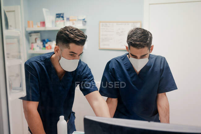 Anónimo jóvenes médicos varones con máscaras respiratorias y uniformes que trabajan en la computadora de escritorio en la clínica - foto de stock