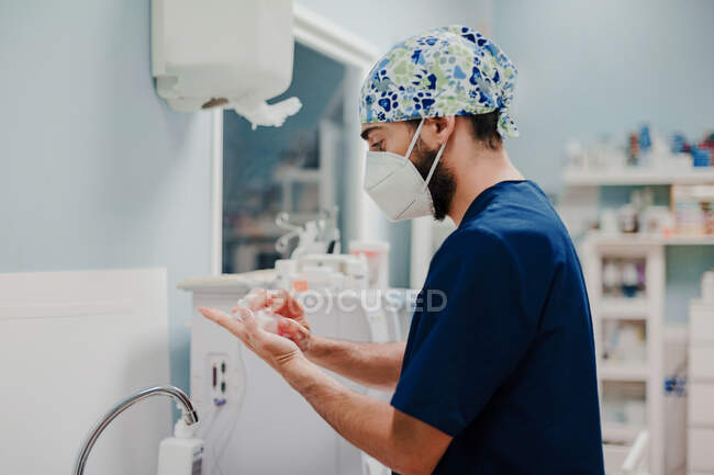 Vue latérale du médecin masculin méconnaissable dans le masque respiratoire et les mains désinfectantes uniformes à l'hôpital — Photo de stock