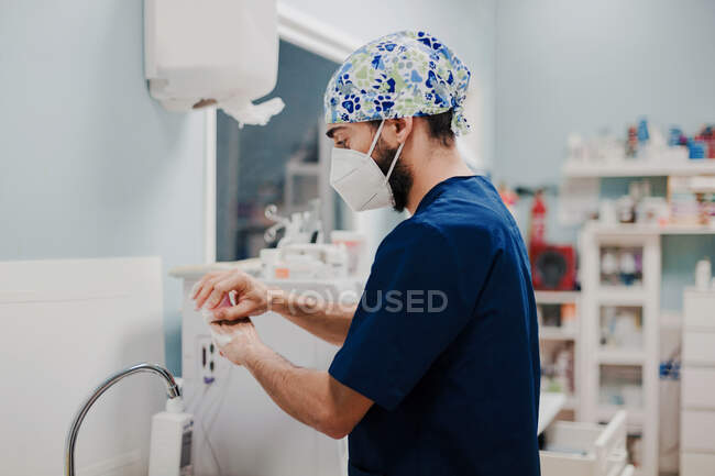 Vista lateral del médico masculino irreconocible en máscara respiratoria y manos desinfectantes uniformes en el hospital - foto de stock