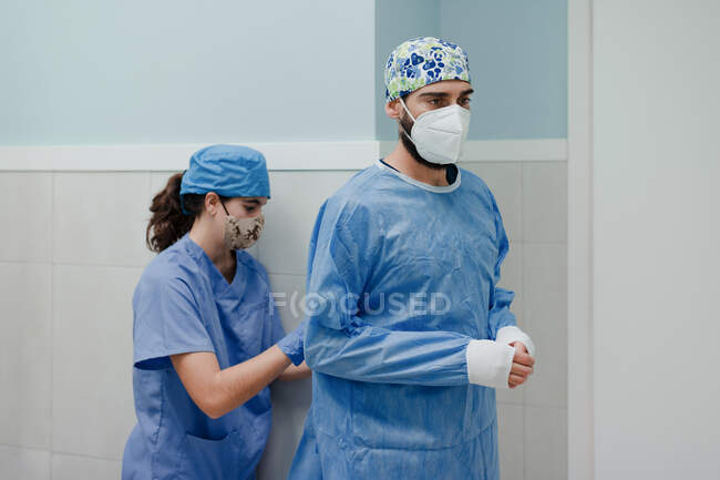 Неузнаваемая медсестра надевает стерильную униформу на мужчину-медика в респираторной маске перед операцией в больнице — стоковое фото