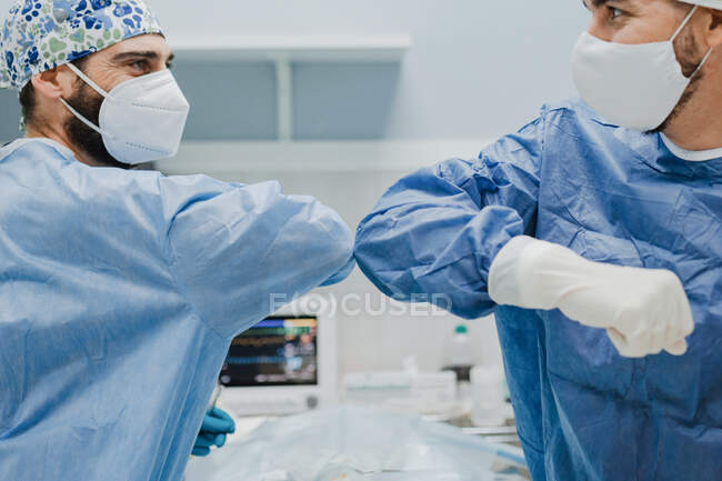Crop anonymous male vet surgeons in respiratory masks looking at each other during greeting before surgery in clinic while bumping elbows — Stock Photo