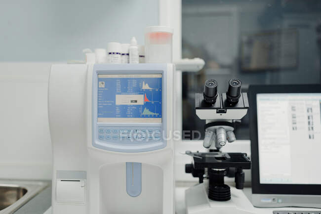 Анализатор гематологии с графиками на мониторе рядом с микроскопом и настольным компьютером с изображениями на экране в лаборатории — стоковое фото