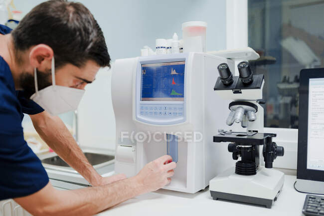 Вид сбоку анонимного врача в респираторной маске с помощью профессионального оборудования с диаграммами на дисплее возле микроскопа в лаборатории — стоковое фото