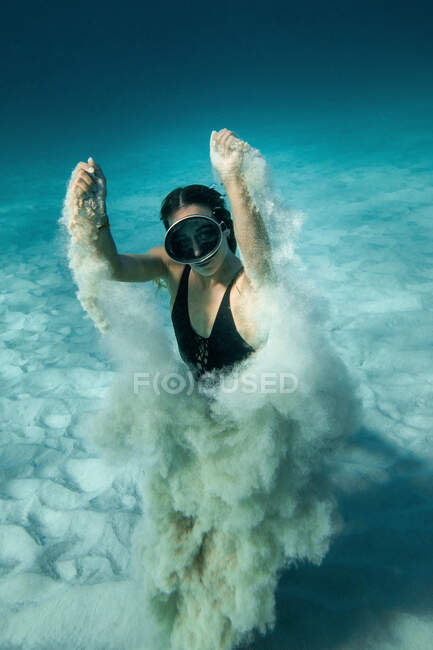 High angle of slim female in goggles and swimsuit swimming underwater and playing with sand while looking at camera — Stock Photo