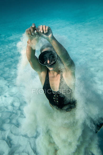 Высокий угол тонкой женщины в очках и купальниках, плавающей под водой и играющей с песком, глядя в камеру — стоковое фото