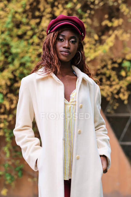 Молодая самоуверенная афроамериканка в модном пальто и берете стоит с руками в карманах, глядя в камеру — стоковое фото