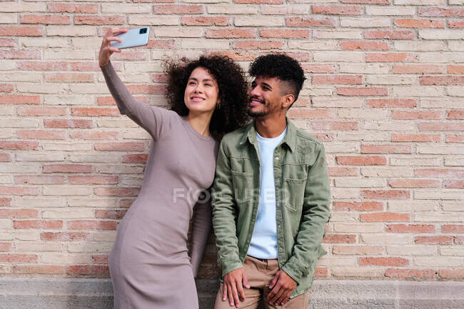 Lächelnde junge multiethnische Freunde in lässiger Kleidung stehen auf der Straße vor einer Ziegelmauer und machen ein Selfie mit modernen Mobiltelefonen — Stockfoto