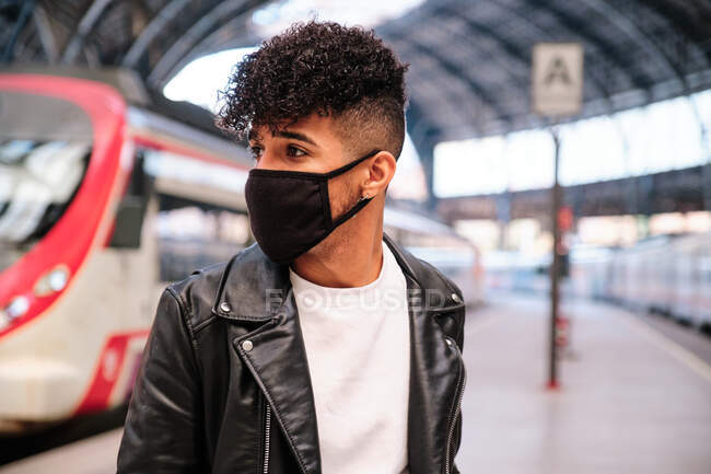 Спокійний молодий етнічний чоловік у шкіряній куртці і захисна маска, стоячи на залізничній платформі і озираючись — стокове фото