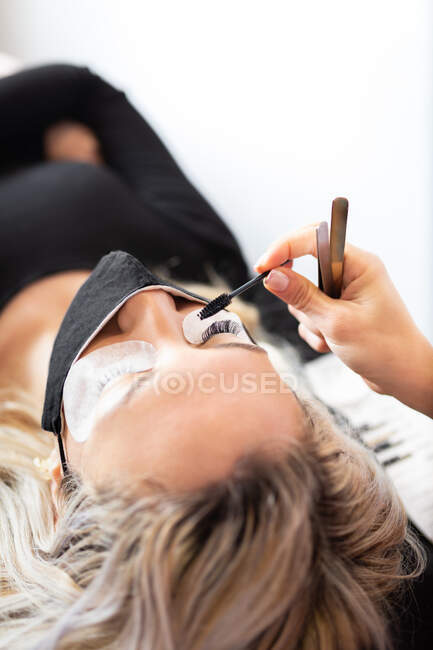 Estetista professionista che applica ciglia artificiali su giovane cliente femminile che indossa maschera protettiva in moderno studio di bellezza — Foto stock