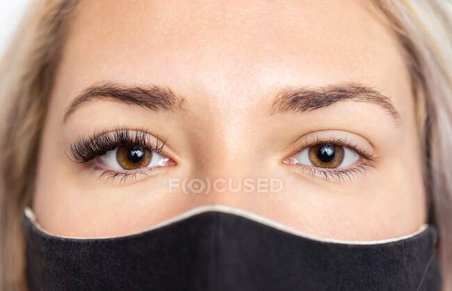 Crop calm young female in black face mask with permanent lashes extension on right eye looking at camera — Stock Photo