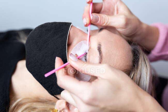 Professionelle Kosmetikerin appliziert künstliche Wimpern auf junge Kundin mit Schutzmaske in modernem Schönheitsstudio — Stockfoto