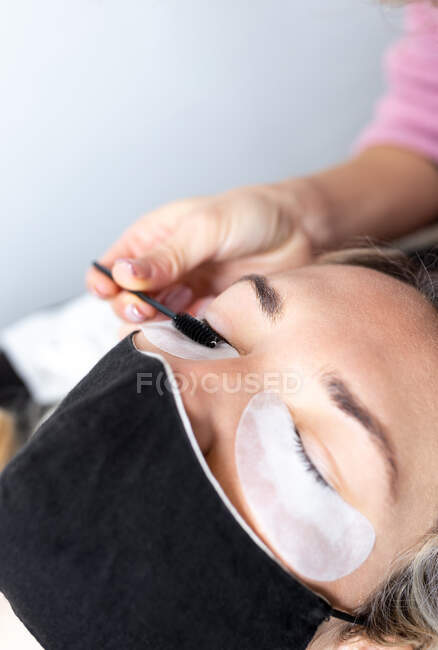Professional beautician applying artificial eyelashes on young female client wearing protective face mask in modern beauty studio — Stock Photo