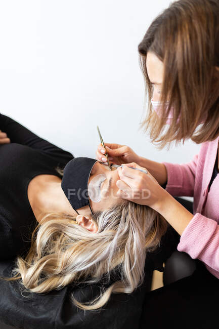 Crop professional female master with tweezer applying artificial eyelashes on young female client in face mask in light modern salon — Stock Photo