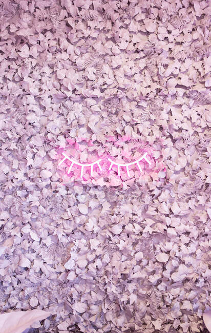 Декоративная креативная стена с цветущими фиолетовыми искусственными цветами и светящимися неоновыми огнями в виде ресниц в современном салоне красоты — стоковое фото