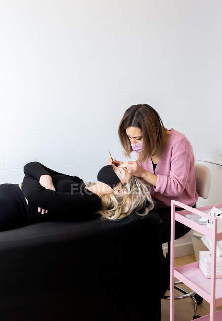 Focused competent female master in casual wear and respirator doing eyelashes extension for slim female customer lying on chair in modern beauty salon — Stock Photo