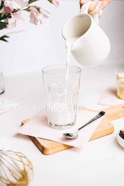 Анонимный урожай, наливающий свежее теплое молоко из кувшина в стекло, помещенное на разделочной доске на светлой кухне — стоковое фото