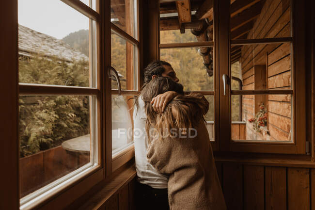 Vista lateral del hombre abrazando novia anónima mientras pasan tiempo juntos en la cabaña de madera a la luz del día - foto de stock