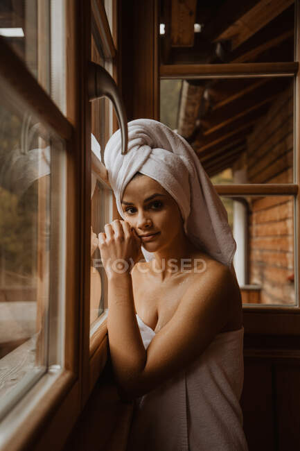 Young gentle female in towels touching face while looking at camera in wooden cabin — Stock Photo