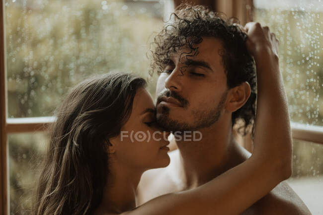 Mujer tierna abrazando suavemente novio barbudo con los ojos cerrados cerca de la ventana con gotas de agua en la cabaña - foto de stock