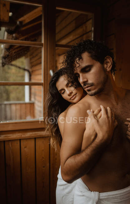 Молодая мечтательная женщина с закрытыми глазами обнимает небритую партнершу с обнаженным туловищем в деревянной хижине — стоковое фото
