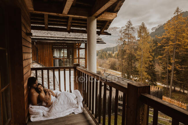 Side view loving young couple bonding tenderly and lying on white soft blanket on wooden cottage terrace on autumn day — Stock Photo