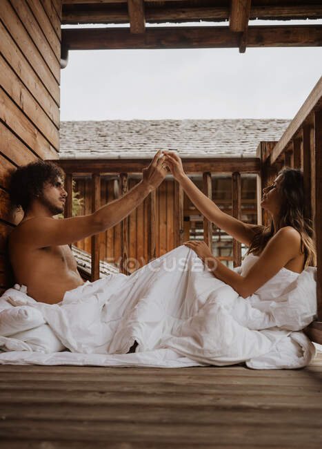 Side view loving young couple touching hands and looking at each other while sitting on terrace in soft white blanket — Stock Photo