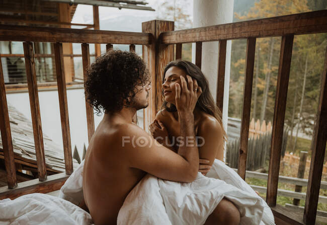 Romântico jovem casal despido abraçando suavemente enquanto sentado no alpendre da casa de campo de madeira e cobrindo corpos com cobertor branco aconchegante no clima de outono — Fotografia de Stock