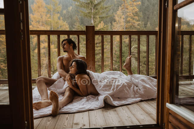 Romántica pareja desnuda tumbada en una acogedora manta y abrazos en la terraza de madera casa de campo contra el bosque de hoja caduca en otoño - foto de stock