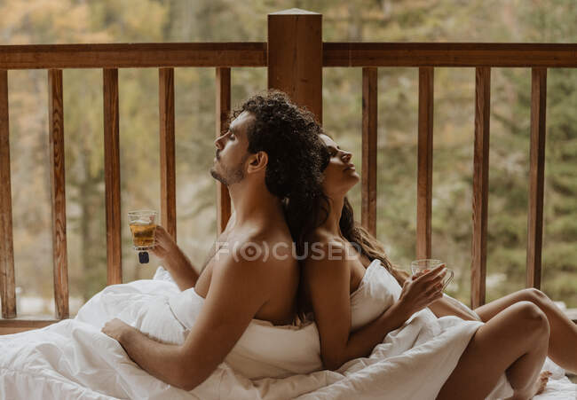 Side view relaxed young undressed couple covering bodies with soft blankets and sitting back to back with eyes closed and cups of teas on terrace on autumn day — Stock Photo