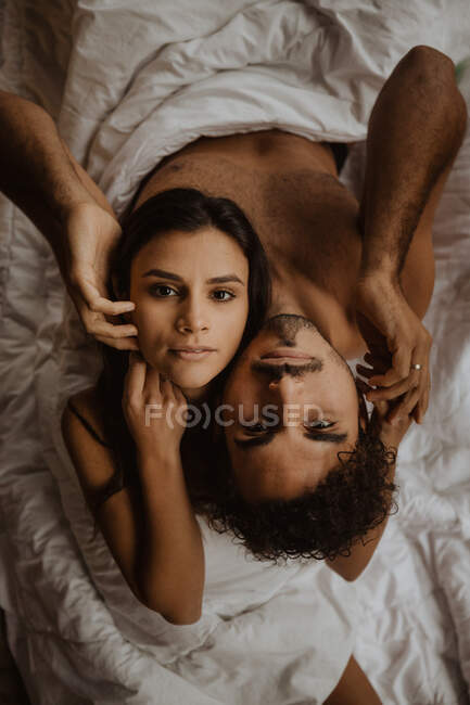 Von oben ein romantisches junges Paar, das Rücken an Rücken sitzt und sich die Köpfe auf die Schultern legt, während es sich entspannt auf einer weichen Decke in die Kamera blickt — Stockfoto