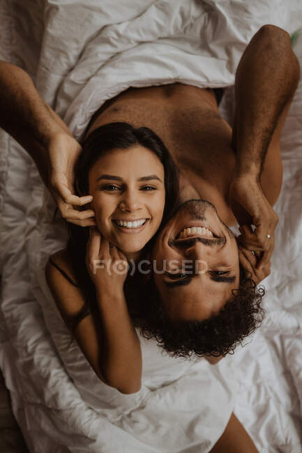 Von oben gut gelauntes unbekleidetes Paar, das Rücken an Rücken auf einer kuscheligen Decke sitzt und mit zahmem Lächeln in die Kamera blickt — Stockfoto