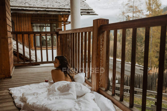 Вид сзади привлекательный топлесс женщина расслабляется на мягкое одеяло с чашкой горячего напитка на деревянном крыльце коттеджа осенью — стоковое фото