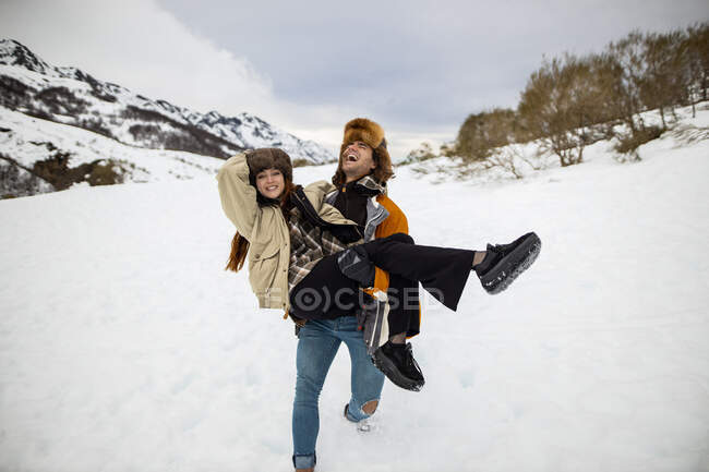 Joven riéndose viajero masculino con los ojos cerrados llevando a la hembra amada en el monte nevado bajo el cielo nublado en España - foto de stock