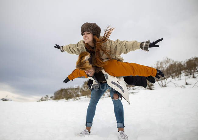 Cheerful male tourist carrying girlfriend with raised arms piggyback on snowy mount in wintertime — Stock Photo