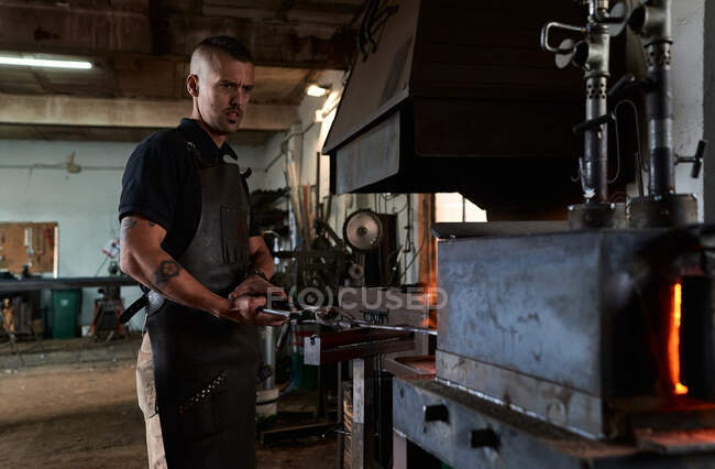 Vue latérale d'un jeune faussaire masculin tatoué grave dans un tablier chauffant du métal dans un four tout en travaillant dans une forge grogneuse — Photo de stock