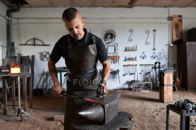 Вид спереди серьезного татуированного молодого мужчины-кузнеца в фартуке, нагревающем металл в печи во время работы в громоздкой кузнице — стоковое фото