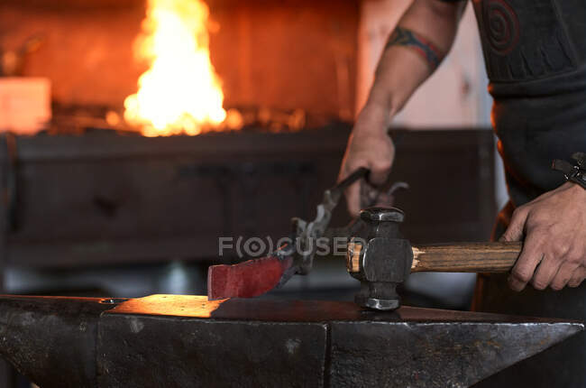 Vista laterale di anonimo tatuato giovane falsario maschio in grembiule riscaldamento metallo in forno mentre si lavora in fucina grungy — Foto stock