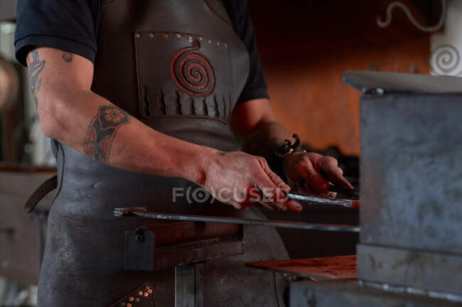 Vista lateral do falsificador macho jovem tatuado anônimo no avental aquecimento de metal no forno enquanto trabalhava em ferreiro grungy — Fotografia de Stock