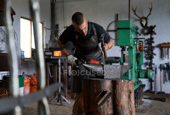 Vista frontal de un joven herrero concentrado con ropa casual y delantal trabajando cerca de yunque con martillo en la mano - foto de stock