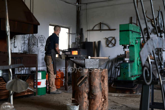 Vue arrière d'un jeune faussaire masculin tatoué grave dans un tablier chauffant du métal dans un four tout en travaillant dans une forge grogneuse — Photo de stock