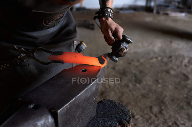 Side view of anonymous tattooed young male forger in apron heating metal in furnace while working in grungy smithy — Stock Photo