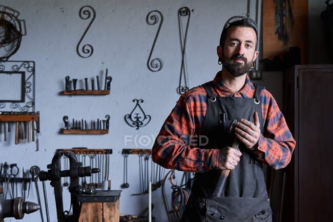 Contenido joven artesano barbudo étnico con camisa a cuadros y delantal sosteniendo el martillo en la mano y mirando a la cámara durante el trabajo en la herrería - foto de stock