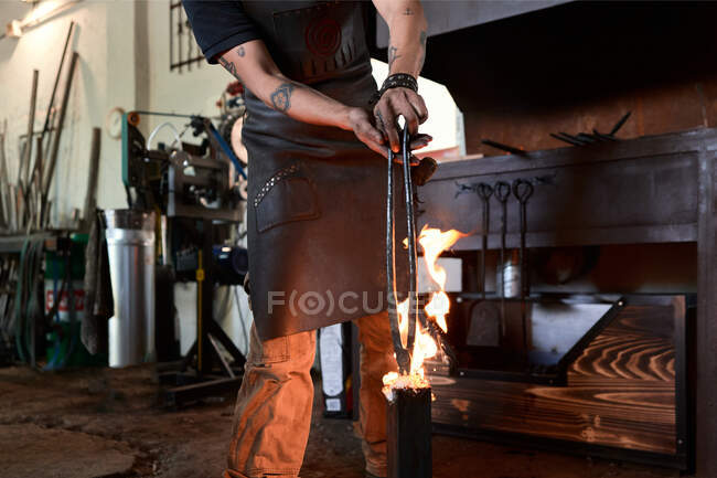 Recadrer forgeron masculin tatoué anonyme dans des vêtements décontractés et tablier chauffant pinces métalliques en flamme pendant le processus de forgeage en atelier — Photo de stock