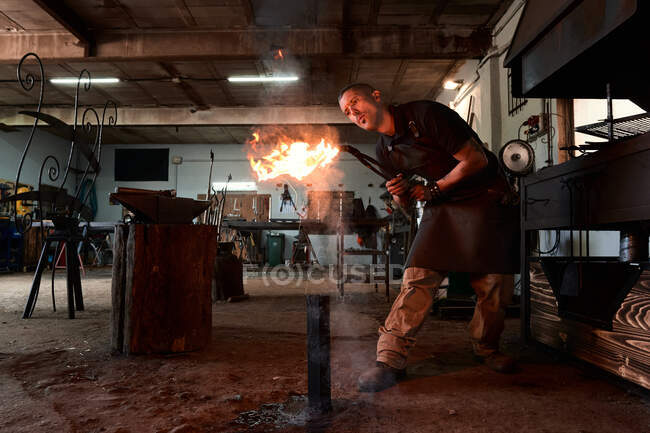 Full body of concentrated young male blacksmith in apron blowing on fire while heating pliers in traditional shabby smithy — Stock Photo