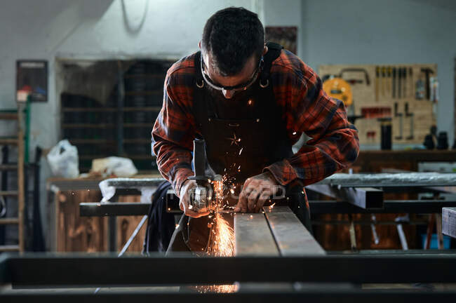 Vue de face d'un jeune forgeron masculin méconnaissable dans des lunettes de protection polissant de petits détails métalliques sur une meuleuse en atelier — Photo de stock