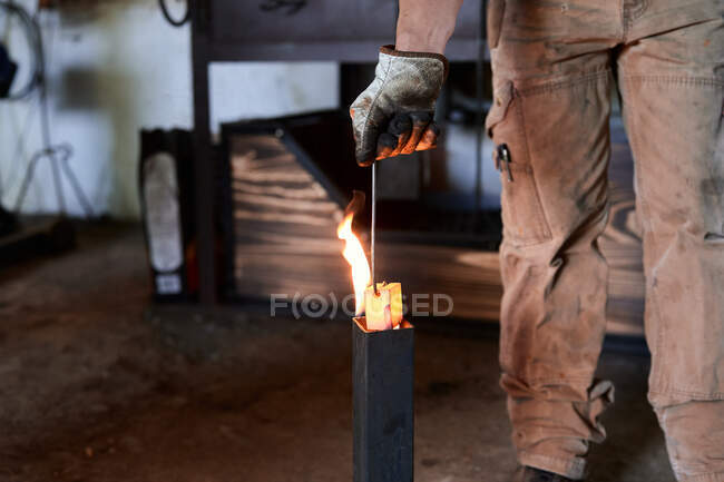 Crop anônimo tatuado ferreiro masculino em roupas casuais e avental de aquecimento de alicates de metal em chamas durante o processo de forjamento em oficina — Fotografia de Stock