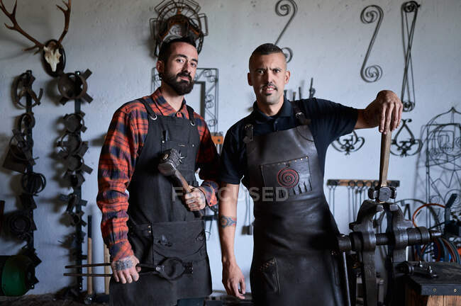 Young brutal ethnic male blacksmiths in casual clothes and aprons standing in workshop with hammers in hands and looking at camera — Stock Photo