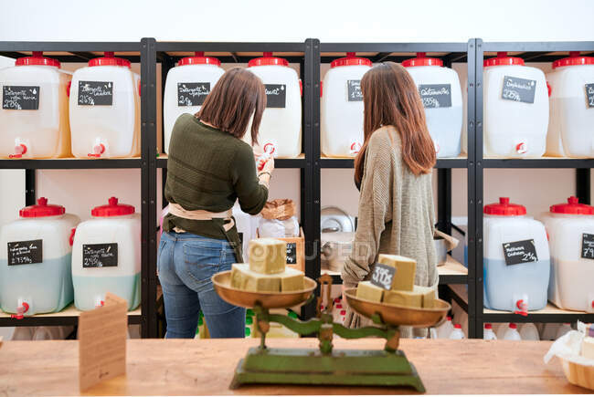 Vista posterior de las mujeres seleccionando el agente de limpieza embotellado colocado en los estantes en la tienda ecológica - foto de stock