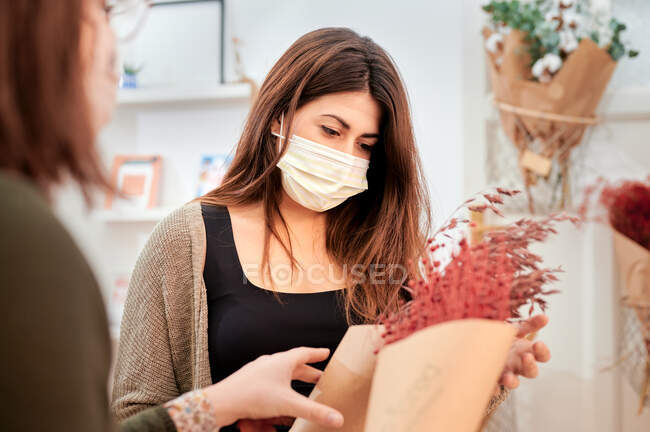 Female customer in protective mask consulting with cropped unrecognizable saleswoman working in store with different bunches of flowers wrapped in zero waste paper package — Stock Photo