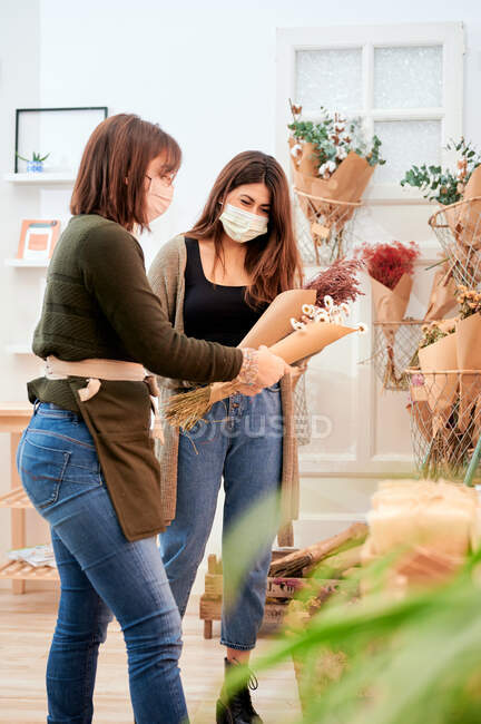 Cliente feminino em consultoria máscara protetora com vendedora trabalhando na loja com diferentes cachos de flores envolvidas em zero pacote de papel residual — Fotografia de Stock